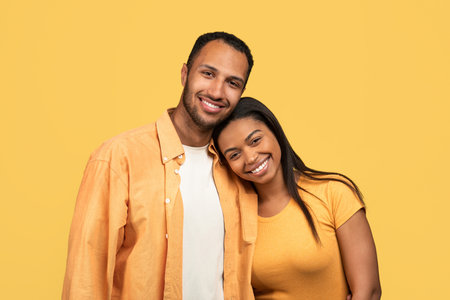 Loving young African American couple cuddling, looking at camera and smiling on yellow studio backgroundの写真素材