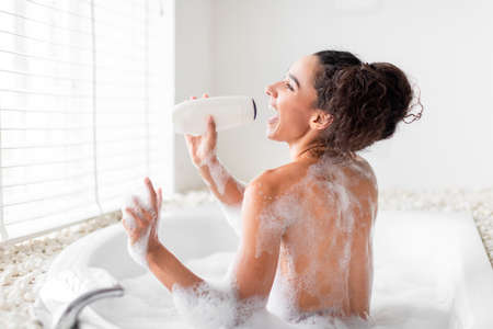 Happy young woman taking bubble bath, using shampoo bottle as microphone, singing and having fun at...