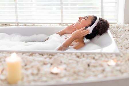 Happy young lady in headphones listening to music while enjoying hot foamy bath at home, blank spaceの写真素材