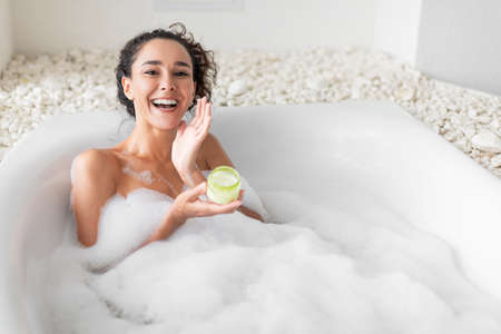 Lovely young woman lying in warm bubble bath, applying cream or face mask, copy space