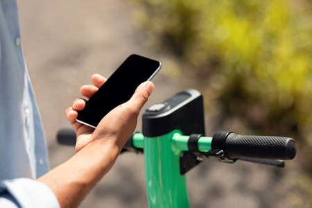 Electro-mobility concept. Black man unlocking e-scooter using his smartphone with black blank screen, mockup for designの写真素材