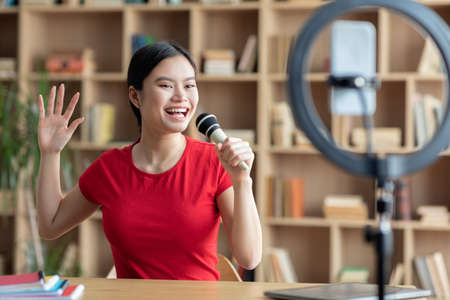 Glad satisfied teen asian woman singing at microphone and shooting video for blog in living room interiorの写真素材