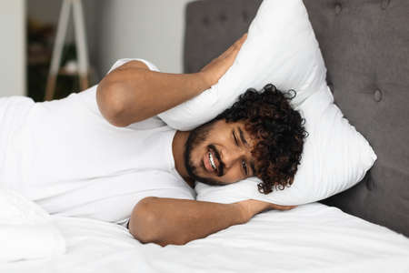 Angry eastern man laying in bed, covering head with pillow