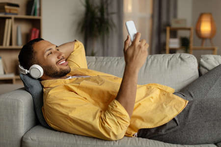 Handsome young black guy in wireless headphones listening to music online on smartphone, lying on couch at homeの写真素材