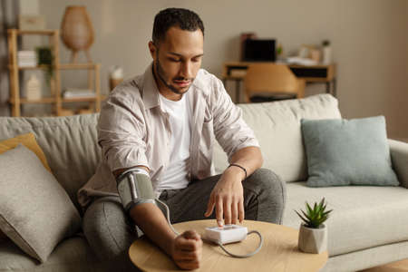 Young African American man measuring arterial blood pressure, having health problem, sitting on sofa at home, copy spaceの写真素材