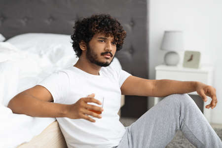 Upset eastern guy drinking alcohol, sitting on floor by bedの写真素材