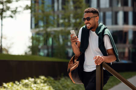 On my way to work. Happy stylish african american man using his smartphone and walking in park area,...