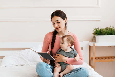 Spending time with baby. Young loving mom reading book to her infant daughter, relaxing together on bed at homeの写真素材