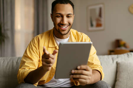 Happy young black man with tablet sitting on couch, working or studying online, having video chat at...