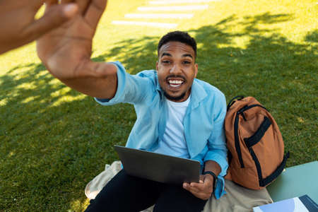 Excited black male student giving high five to friend while using laptop computer, sitting on grass...