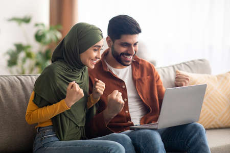 Cheerful Muslim Couple Using Laptop And Gesturing Yes At Homeの写真素材