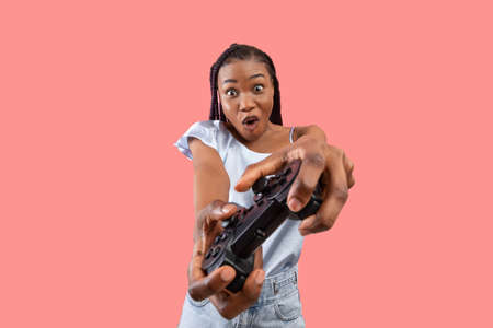 Excited young black lady with gaming controller playing video game on pink studio background