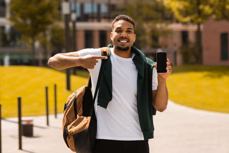 Great offer. Happy black guy standing outdoors, holding cellphone and pointing at phone blank screen