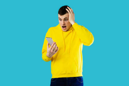 Shocked Millennial Guy Holding Smartphone Reading Negative Message, Blue Background