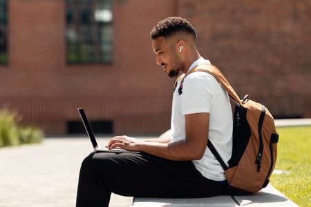 African american freelance man sitting outdoors in park, typing on laptop while working online with computer, side viewの写真素材