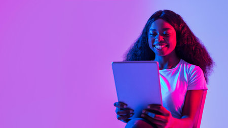 Portrait of joyful young black woman using tablet pc, studying or working remotely, having online meeting in neon lightの写真素材