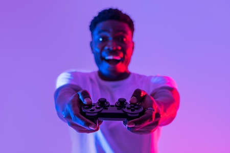 Happy young black guy with joystick playing video game in neon light, selective focus. Modern hobbiesの写真素材