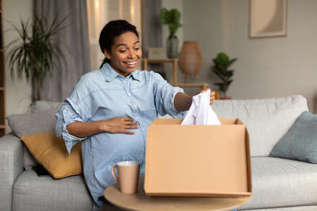 Excited Pregnant Black Lady Unpacking Box With Clothes Sitting Indoor