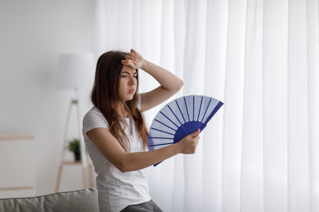 Despaired tired millennial caucasian lady suffering from heat waves blowing fan at herself, wipes sweat with handの写真素材