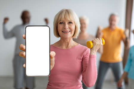Happy senior woman with dumbbell in hand showing big blank smartphone