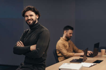 Attractive young businessman posing with crossed arms and smiling at camera, his coworker using...
