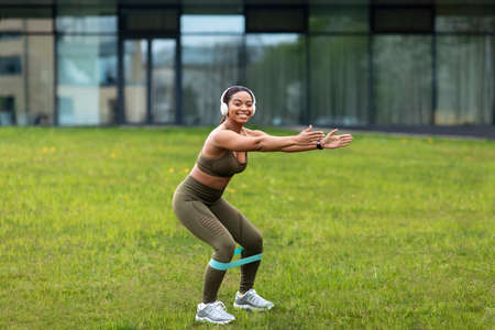 Sporty young African American woman doing squats with elastic band, listening to music in headphones...