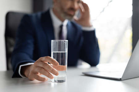 Stressed businessman suffering from headache at workplace, holding glass of waterの写真素材