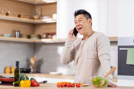 Positive chinese man cooking healthy salad and talking on phoneの写真素材
