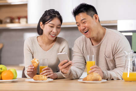 Happy asian couple using cell phone while having breakfastの写真素材