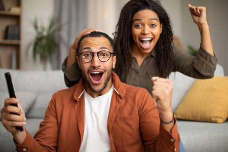 Joyful Black Couple Shaking Fists Watching Sport On TV Indoorの写真素材