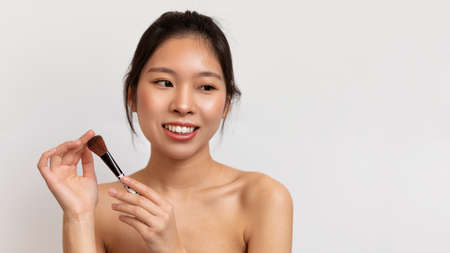 Daily makeup concept. Portrait of young asian lady with natural beauty holding blusher brush, posing on white backgroundの写真素材