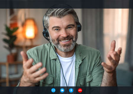 Smiling senior european man with beard in headphones gesturing, looks at camera on screen with buttonsの写真素材