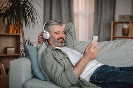 Happy mature european male in headphones watch video at smartphone, lies on sofa and rests in living...