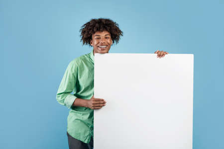 Happy black teen guy holding empty paper poster with mockup for your ad design on blue studio backgroundの写真素材