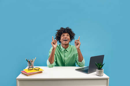 Happy black guy student pointing up at copy space, sitting at desk with books and laptop on blue...