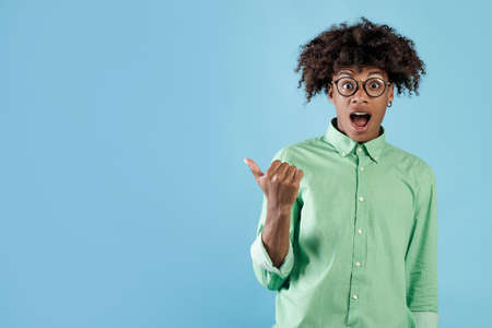 Emotional african american guy standing with open mouth and pointing at free space on blue studio backgroundの写真素材