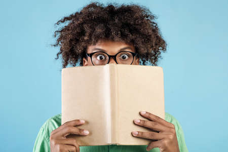 Portrait of emotional african american guy in glasses covering face with book, hiding behind it with...