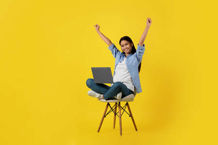 Satisfied excited young korean female sit on chair with laptop, raising her hands, making success gestureの写真素材