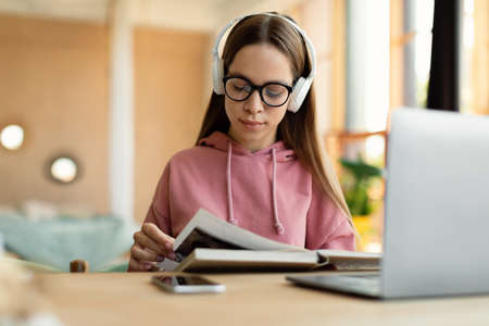 Smart teen girl reading textbook, sitting at table with laptop and studying materials, learning...