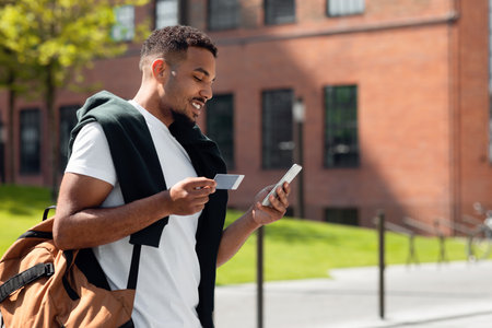 Stylish african american man using mobile phone and holding credit card, purchasing or making online payment outdoorsの写真素材