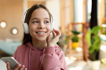 Happy teen girl in headphones using mobile phone, enjoying music or listening audio book, sitting at...