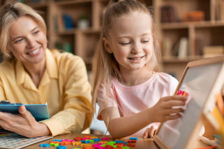 Friendly woman teaching adorable girl alphabet, making words from colorful plastic letters on board