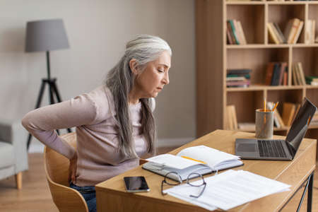 Unhappy caucasian old female with gray hair works on computer and suffers from back pain in room...