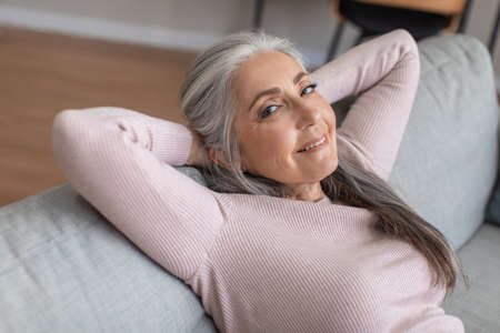 Smiling european old woman with gray hair resting in spare time, enjoy comfort and look at camera in room interiorの写真素材