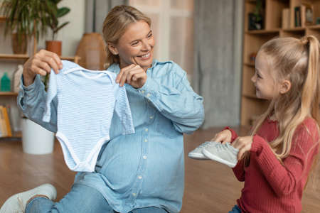 Happy pregnant woman showing clothes for her baby, expectant mother with her elder daughter sitting on floor at homeの写真素材
