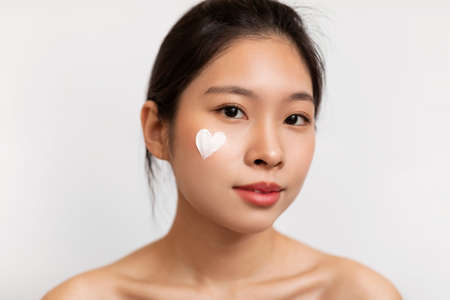 Closeup portrait of pretty korean lady with moisturizer cream on her face in heart shape, white studio backgroundの写真素材