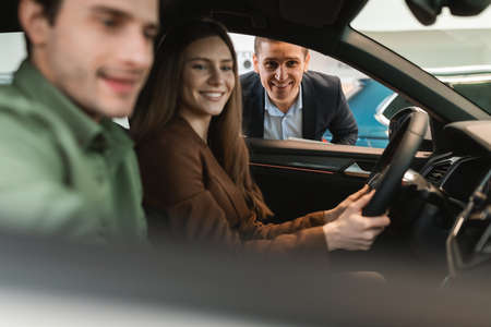 Young couple buying new car, sitting in auto salon, going on test drive, talking to salesman at...