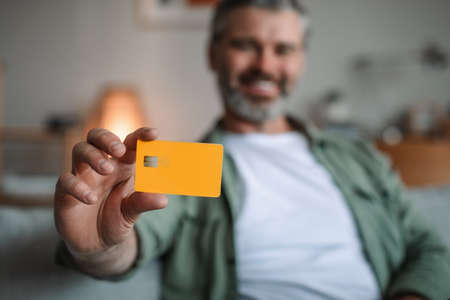 Smiling caucasian old man with beard shows credit card for finances in living room interior, selective focusの写真素材