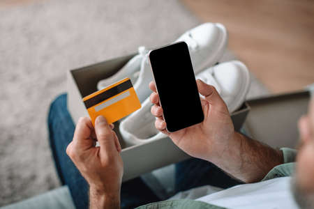 Caucasian old man with cardboard box with sneakers uses credit card and smartphone with empty screen
