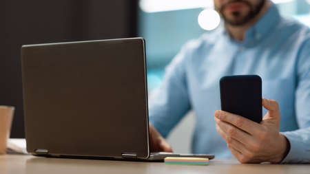 Unrecognizable mature businessman typing on smartphone, using laptop at office, selective focus....